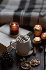 Aromatherapy on a grey fall morning, atmosphere of cosiness and relax. Autumn cozy home composition with hot chocolate with marshmallow and candles. Wooden background, books, close up.