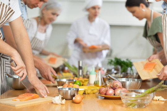 People in aprons standing at table with groceries and utensils during group cooking lesson, absorbed in cooking process, cutting fresh salmon following chef instructions. Cropped shot