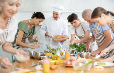 Positive middle-aged male cook conducting culinary classes for learners standing around table with...