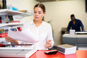 Concentrated young woman in white shirt using paper cutter on the table with planners and calculator in the printer house