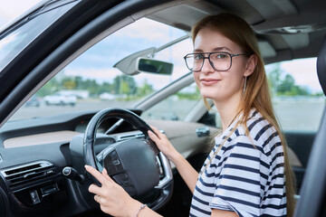 Fototapeta na wymiar Teenage girl driver in glasses sitting behind wheel of car, looking at camera