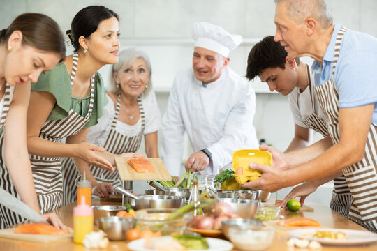Friendly young Asian woman holding cutting board with fresh salmon steak and talking to elderly man during group culinary classes..
