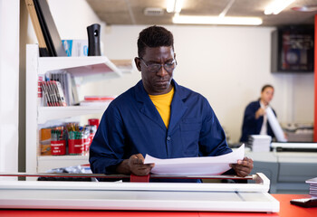 Serious middle-aged African American male in a blue uniform using paper cutter in the printer house