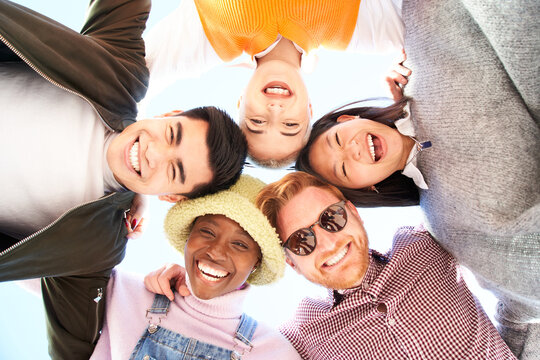 Multiracial and divers group of friends hugging in circle and looking down at the camera. They stand happy outside. Putting their heads together. Low angle of a young people taking selfie