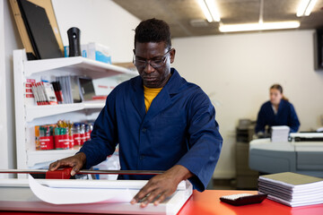 Fototapeta premium Focused African American man in blue robe uniform trimming the printed work on the cutter in the typography