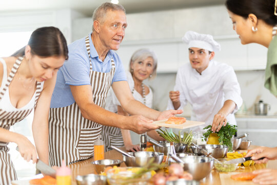 Enthusiastic senior man honing culinary skills during group master class from professional chef, holding wooden cutting board with fresh salmon steak and talking friendly with other participants.. - Powered by Adobe