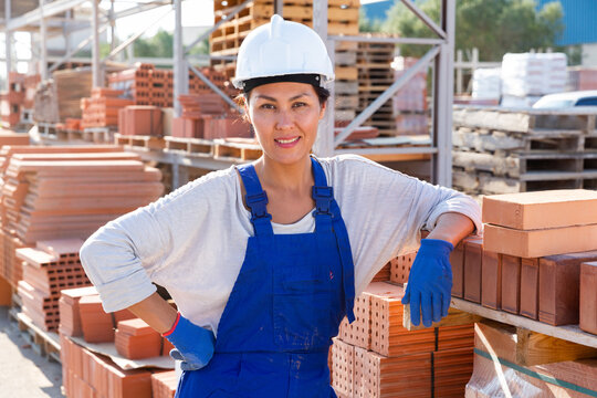 Asian female builder standing amongst brick stacks in outdoor construction material storage.