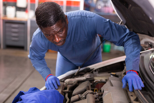 Afro american car mechanic looking at open hood for internal checking of vehicle in workshop