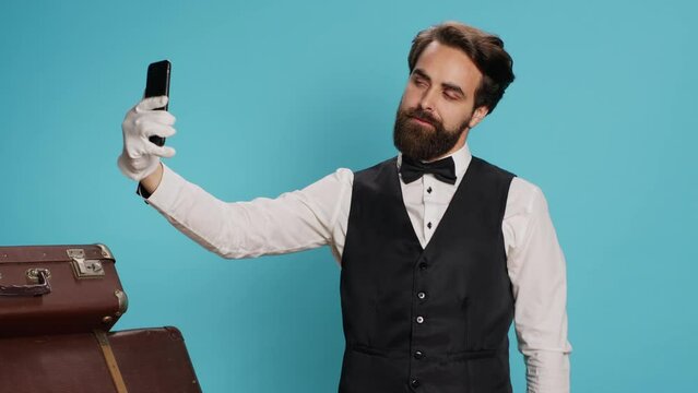 In studio, doorman in uniform takes photos while using smartphone for selfies and acting silly against blue background. Skilled hotel concierge does pictures with his gadget while on the job.