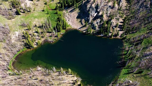 Drone Aerial Shot Pulls Out and Pans Up on a Backcountry Backpacking Lake. Surrounded by forest and towering stone rock mountains. Patches of snow are scattered along the ridge top.
