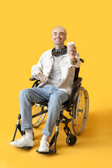 Young tattooed man in wheelchair with cup of coffee on yellow background