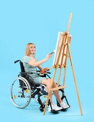 Young woman in wheelchair painting on easel against blue background