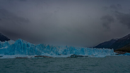Naklejka premium A wall of blue ice with sharp spikes rises above a turquoise lake. Cloudy. Mountains in the fog. Icebergs float in the water. Perito Moreno glacier. Los Glaciares National Park. El Calafate. Argentina