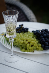 A dish with grapes and a crystal glass stands on a white table, in the open air, in a summer cottage.