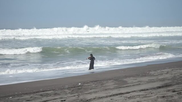 A woman in black dress playing violin on the beach facing the ocean waves
