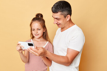 Smiling satisfied man and little kid girl standing together isolated over beige background using mobile phone together  playing games or watching cartoons spending time together