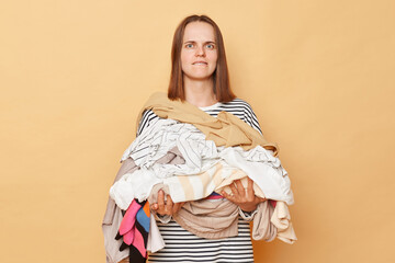 Confused brunette woman holding pile of clothes in hands isolated over beige background looking at...