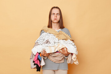 Tired exhausted brunette woman holding pile of clothes in hands isolated over beige background...