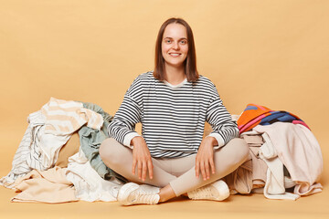 Smiling cheerful attractive Caucasian woman posing near heap of multicolored unsorted clothes...