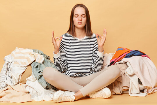 Calm relaxed Caucasian woman posing near heap of multicolored unsorted clothes isolated over beige background meditating practicing yoga has lots of household work.