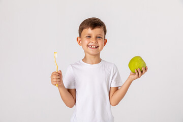 Little boy in white t-shirt smiling, holding in his hand toothbrush and green apple on white studio...