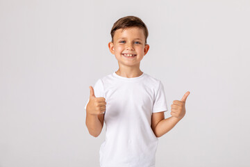 Happy smiling satisfied caucasian boy shows thumbs up looking at camera, portrait. attractive smiling boy in white casual shirt posing on white background. childhood, human emotions, lifestyle - obrazy, fototapety, plakaty