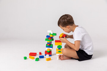 Toddler playing with educational blocks. Side view happy child collects a tower of colored cubes. The preschooler sits down at the white background and assembles the construction set.