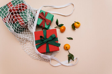 Gifts with ribbons are in a white eco-friendly string bag on a beige background, next to orange ripe tangerines with leaves. Top view. Copy space