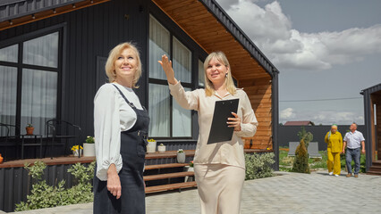 Blonde realtor talking to woman ready to buy summer house for elderly parents. Potential customer listens to real estate agent introducing offered cottage