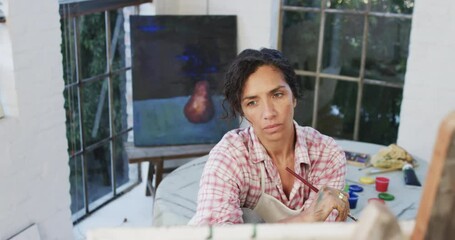 Biracial female artist in apron holding brush and looking at canvas in studio, slow motion