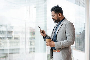 A professional Indian-Asian businessman is checking messages on his phone while standing in a building.