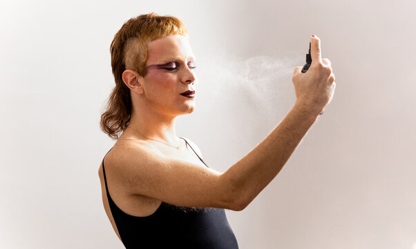 A non-binary redhead with makeup poses with their eyes closed on a white background while using a spray to set makeup. Concept of non-binary people, makeup fixer. Men in makeup.