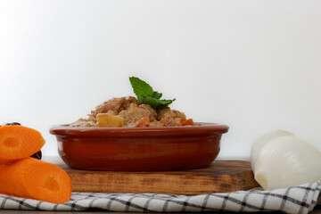 meat stew with sauce in a clay pot on a wooden board accompanied by carrots and onions