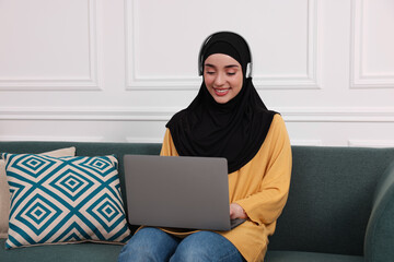 Muslim woman in hijab and headphones using laptop on sofa indoors