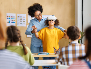 Innovation in education. Student schoolgirl in virtual reality glasses together with teacher during lesson in classroom at school - 638350981