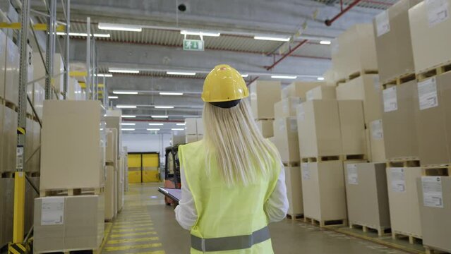 Blonde woman employee in protective vest and helmet walking in warehouse back view handheld tracking shot. Storehouse worker checking stock management in storage