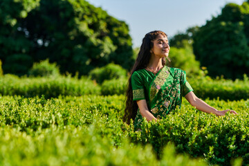 Naklejka na ściany i meble pleased young indian woman in sari standing with closed eyes near green plants in park