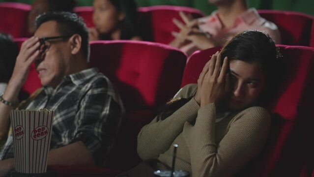 Asian man and woman having shocked and frightened reaction while watching horror thriller movie in the movie theater. Scared woman and man covering eyes when watching horror movie