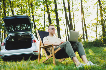Obraz premium A cheerful man in glasses sits on a chair in nature and works on vacation, work online on a portable laptop. Freelance work.