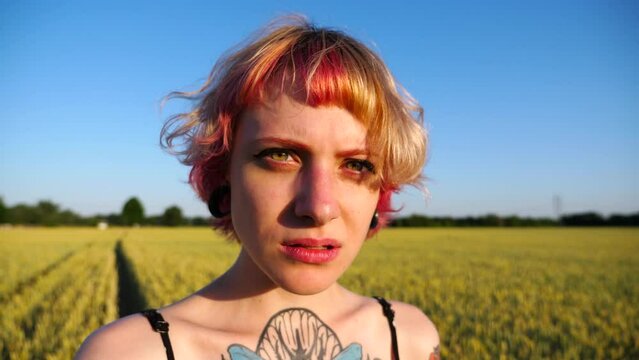 Portrait of serious punk girl with ear tunnels with blurred wheat green field at background. Unhappy hippie woman with pink hair looks into camera standing at barley meadow. Close up