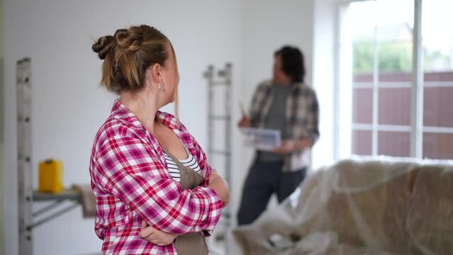Satisfied woman on the left looking back at blurred man and looking at camera in slow motion. Portrait of confident Caucasian happy wife posing repairing house with husband indoors