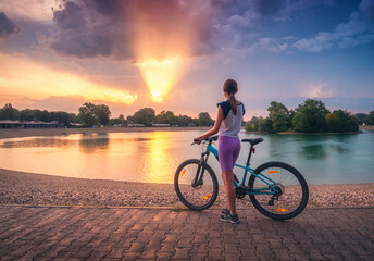 Woman riding a mountain bike near lake at sunset in summer. Colorful landscape with sporty girl riding a bicycle, beach, beautiful sky reflected in water in park in spring. Sport and travel. Biking