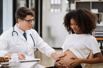 Fototapeta premium Happy pregnant woman has appointment with doctor at clinic. male medic specialist with stethoscope listens to baby's heartbeat in mother's belly. Pregnancy, health care concept.