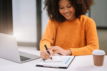 Business woman using laptop to work and writing on notebook with pen in office with mobile next to laptop.