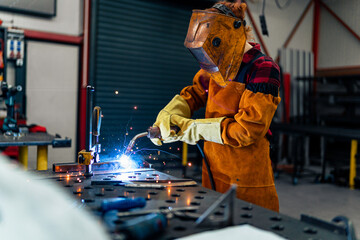 A woman in protective equipment uses tools and machines in the workshop, sparks fly and illuminate...