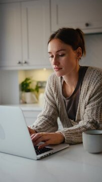 Vertical video of young woman working from home on laptop standing by kitchen counter - shot in slow motion