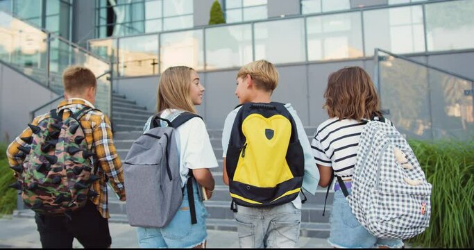 Rear view. Good-looking school child friends climb stairs going to class with backpacks. Boys and girls high school students having fun after school. They are smiling and having fun.