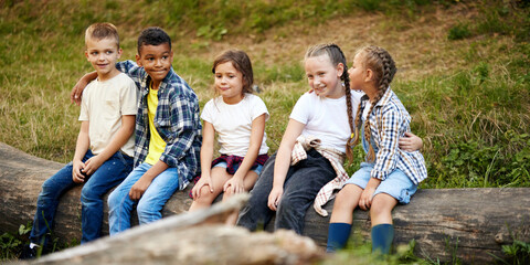 Cheerful playful kids sitting on log in forest on sunny day. Going hiking, walking, having active weekends. Concept of leisure activity, childhood, summer, friendship, active lifestyle, fun, nature