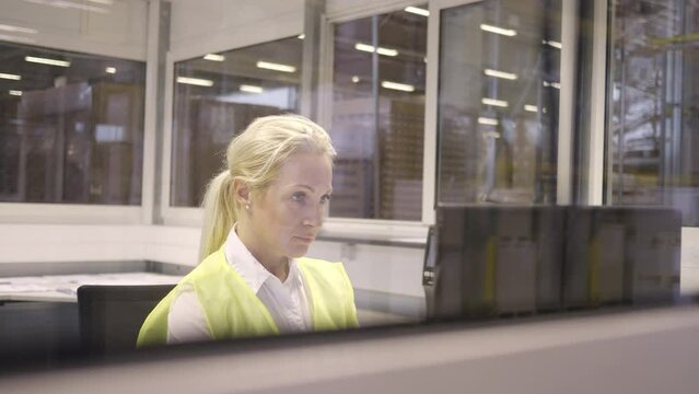 Blonde mid adult woman working on computer in warehouse office behind window. Shipment manager planning goods distribution with software in storehouse