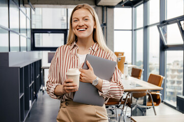 A young girl is standing with a coffee and a laptop in her hands in a modern work area